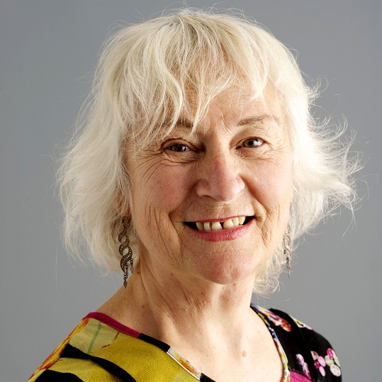 Colour photograph of Bonita Ely, head and shoulder shot, Bonita wears a abstract top with shapes and colours, gold, yellow, magenta, black, white, blue, pink and green. Bonita has an open mouth smile and is wearing long silver flat swirling earrings, she has white hair cut above her shoulders 